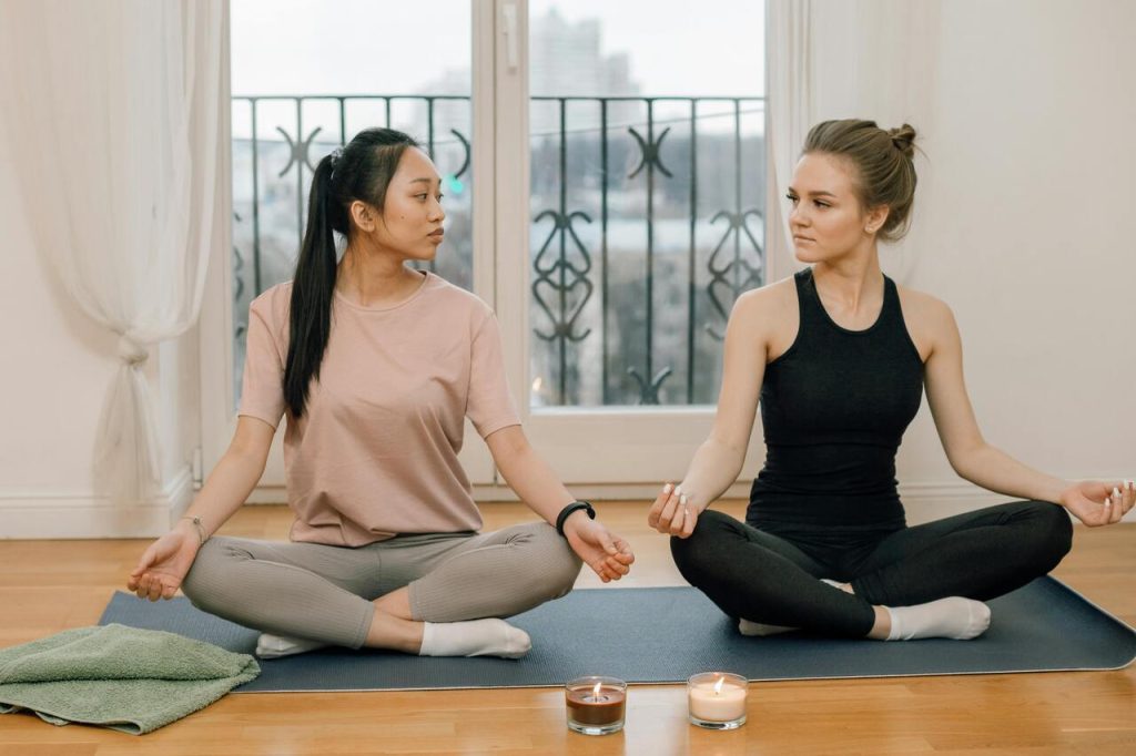 Duas mulheres sentadas num tapete de ioga, em posição de meditação. Elas de frente uma para a outra, com as mãos reforçadas nos joelhos e os olhos fechados ou próximos abertos. O ambiente é tranquilo, com velas acesas no chão e uma vista para uma varanda ao fundo.