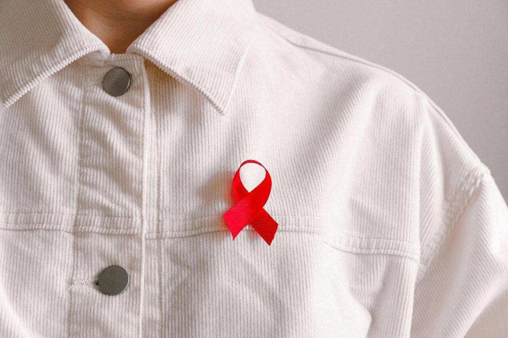 A imagem mostra uma pessoa vestida com uma camisa branca de bombazina, com um laço vermelho preso ao peito. O laço vermelho é um símbolo internacional da luta contra a VIH/SIDA e da consciencialização sobre esta doença.