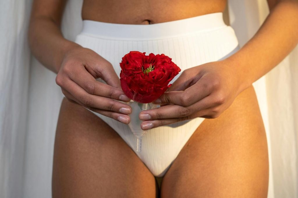 mulher segurando um copo menstrual com uma flor vermelha, simbolizando a menstruação e a saúde íntima feminina. Sugere um tom de naturalidade e empoderamento em relação ao corpo e ao ciclo menstrual